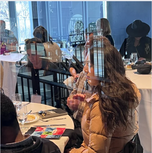 People at a dining table. Reflections of building visible in window. Woman in tan jacket, others seated.