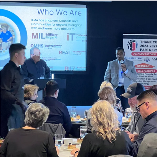 People at a conference with a speaker, presentation, and sponsors.