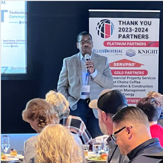 A man in a suit speaks into a microphone at a business event. A banner lists partners.