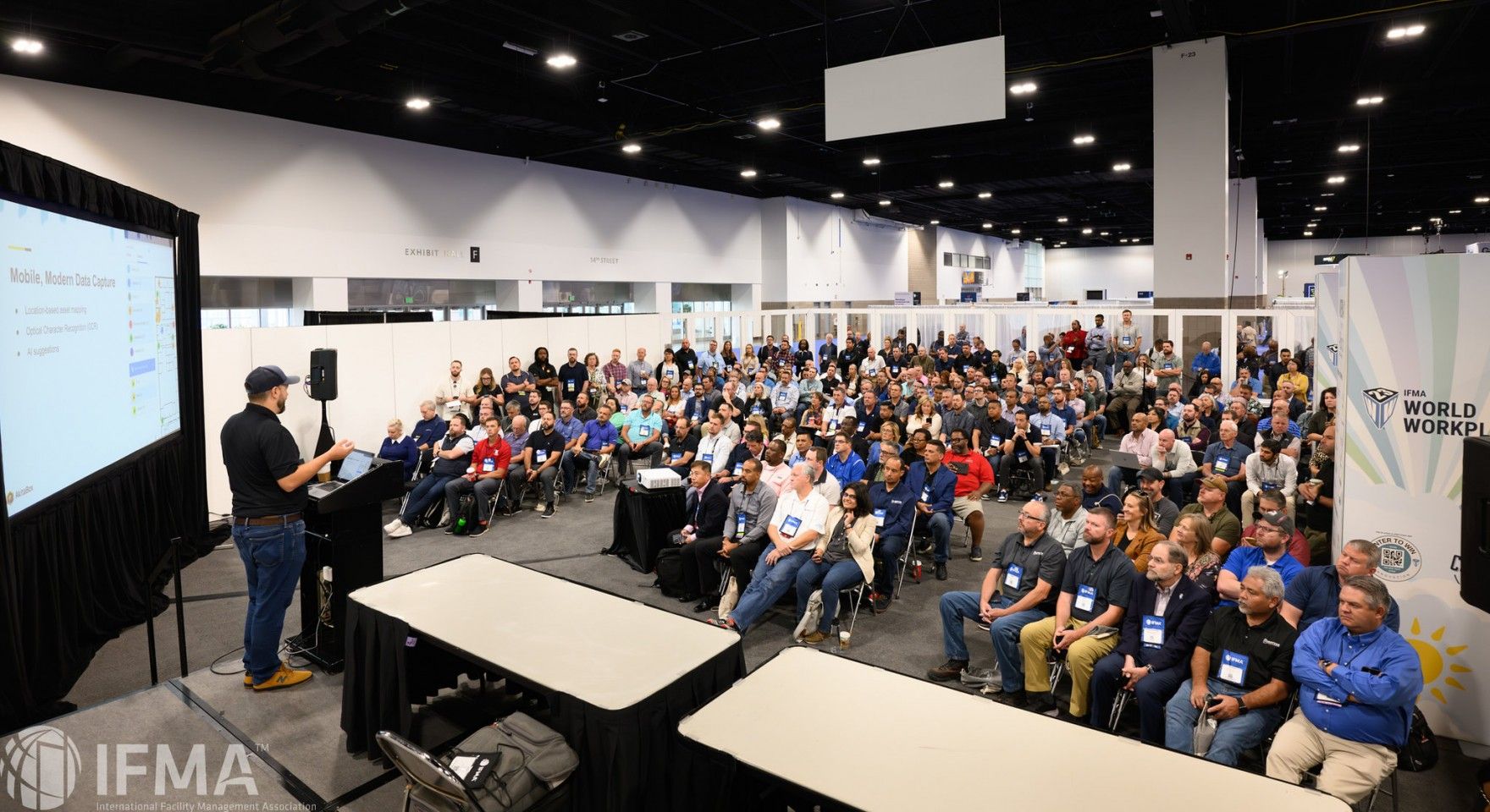 Man presenting to a large audience at a conference. The room is brightly lit with many attendees seated.