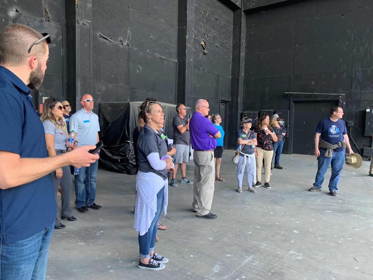 A group of people standing inside a large, black-walled space, possibly a theater. Some are looking towards the right.