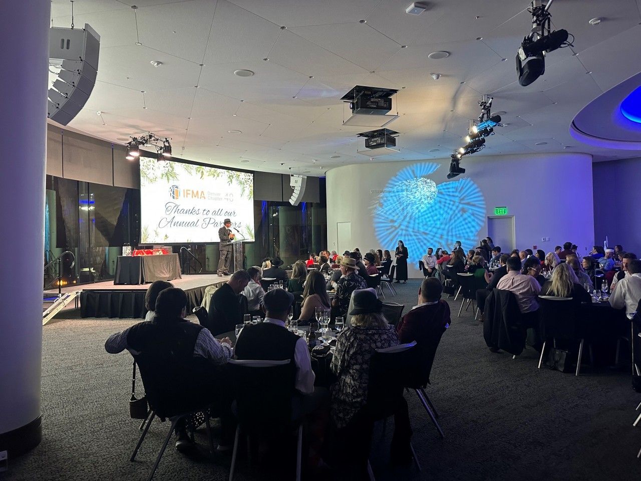 People seated at tables in a banquet hall, watching a presentation on stage.