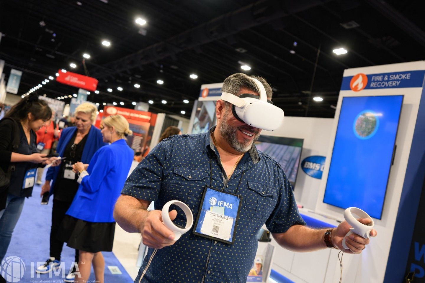 Man using VR headset at trade show, holding controllers, blue shirt. Others look on.