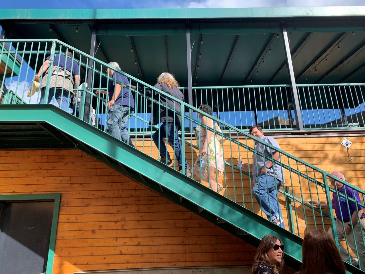 People ascending a teal metal staircase to a second story; orange building exterior, blue sky visible.