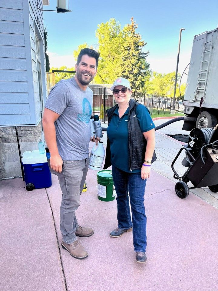 Man and woman smiling, standing outside near a truck and a building. They hold containers. Sunny day.