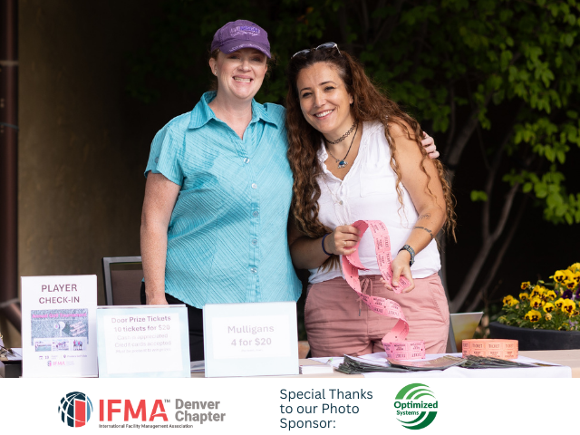 Two women smiling at a table, one in blue, one in white with a pink ribbon. Outdoor event. IFMA logo.