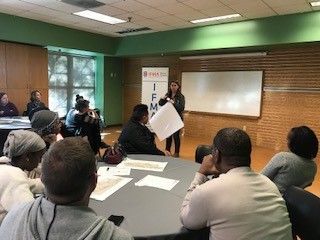 A woman presents a paper to a group seated around tables in a room with a whiteboard.