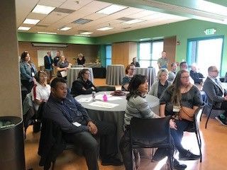 People seated at tables in a room, some looking at a speaker. A few stand near the back.