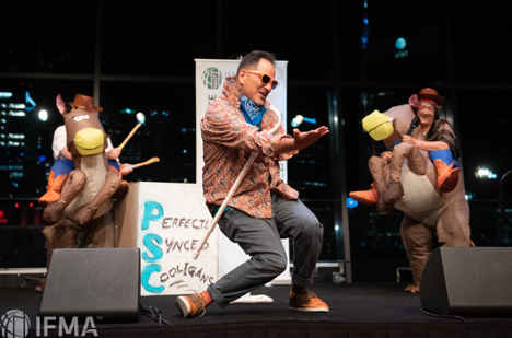 Man dancing on stage with two people in horse costumes. Night backdrop, stage sign.