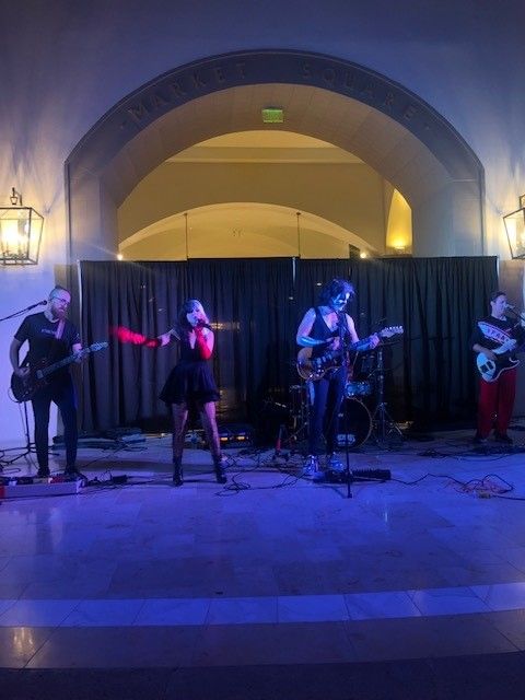 Band performing on stage under an arch. Singer in black dress, red gloves; others play guitars. Blue lighting.