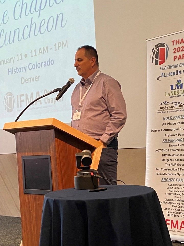 Man speaking at a podium with a microphone at a luncheon in Denver.  Event banner in background.