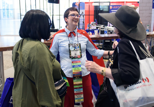Three people talking at an event. A person in a superhero costume smiles. Two other people stand near them.