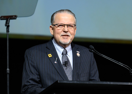 Man in suit speaking at a podium, smiling. A microphone and stage are visible.