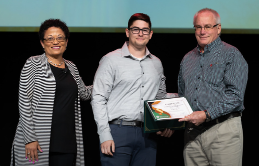 Three people on a stage: a man receiving a certificate from another man, with a woman standing nearby.