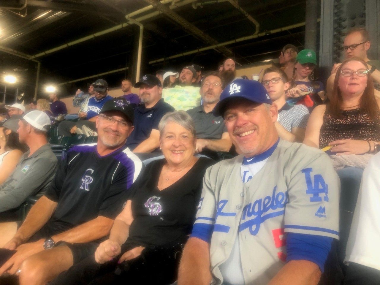 People in baseball stadium seats; two men and a woman smiling, wearing sports attire; others in background.
