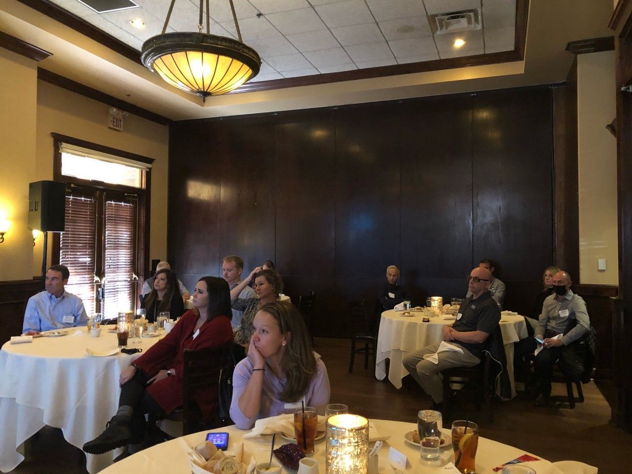 People seated at round tables in a restaurant, listening to a presentation. Dim lighting, dark wood paneling.
