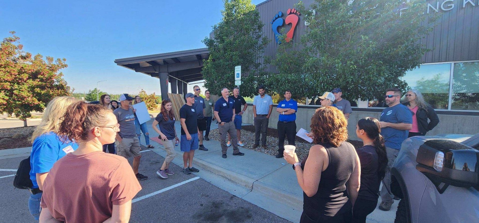 Group of people outside a building with a colorful logo. They appear to be listening to a speaker. Blue sky.