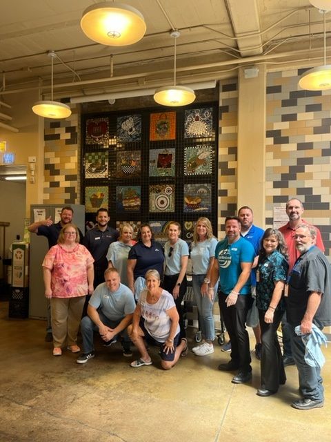 Group of people posing indoors in front of a tile mural. Well lit room with neutral colored walls and ceiling.