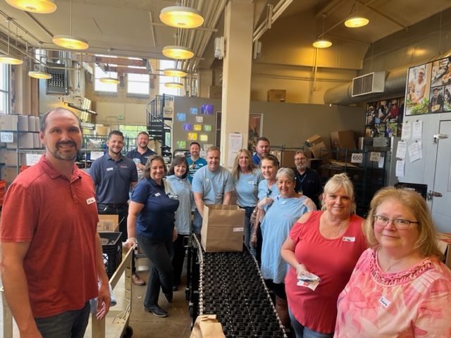 Group of people by a conveyor belt, some holding a brown paper bag. Inside a warehouse-like setting with boxes and supplies.