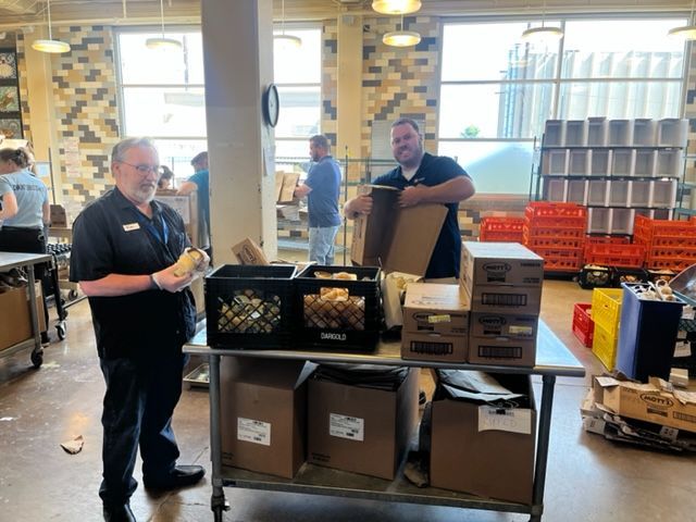 Two men packing boxes at a food bank, other volunteers in background. Bright interior with shelves.