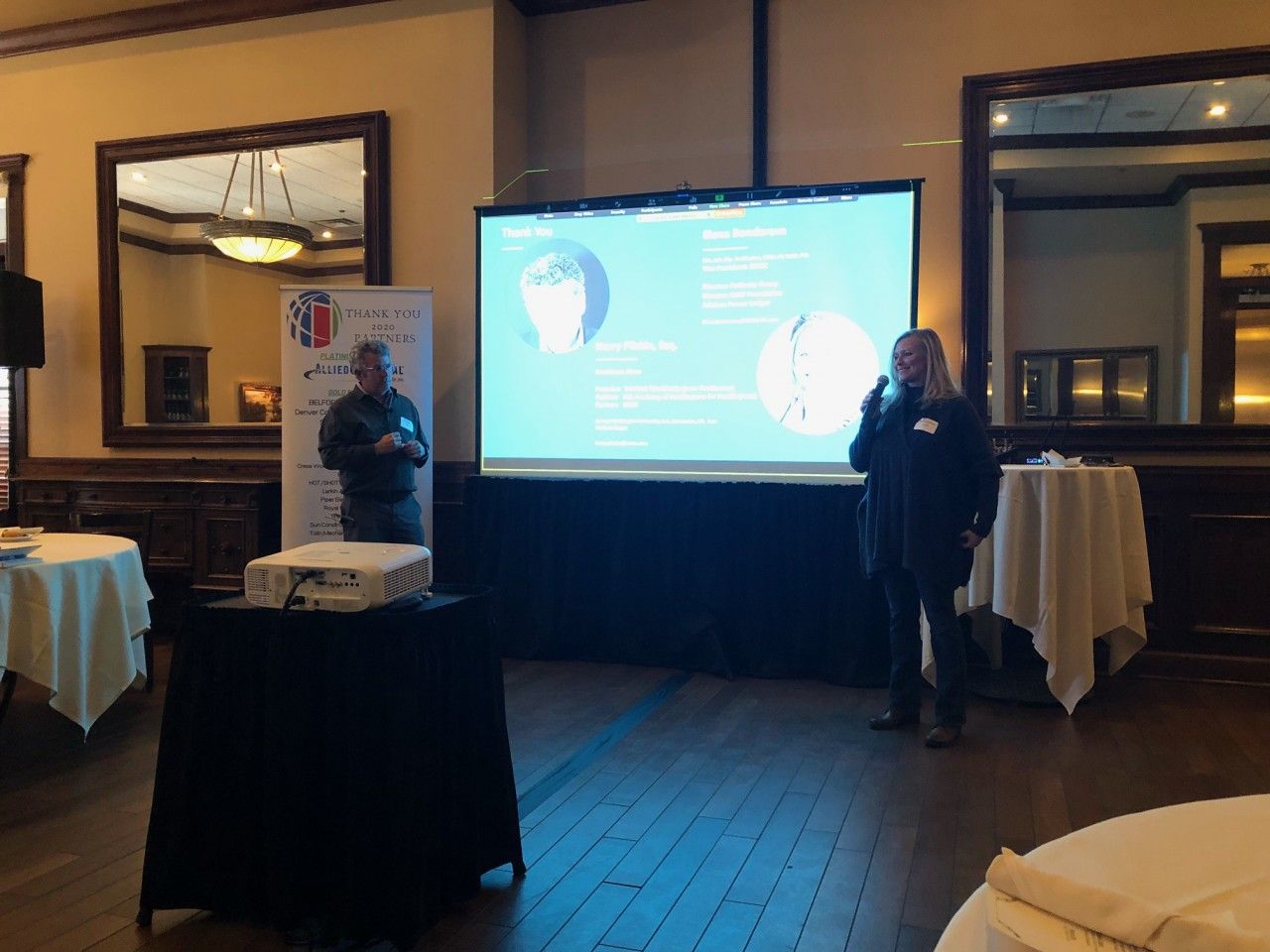 Two people presenting in a room, with a projected screen. Tables with white tablecloths are in the foreground.
