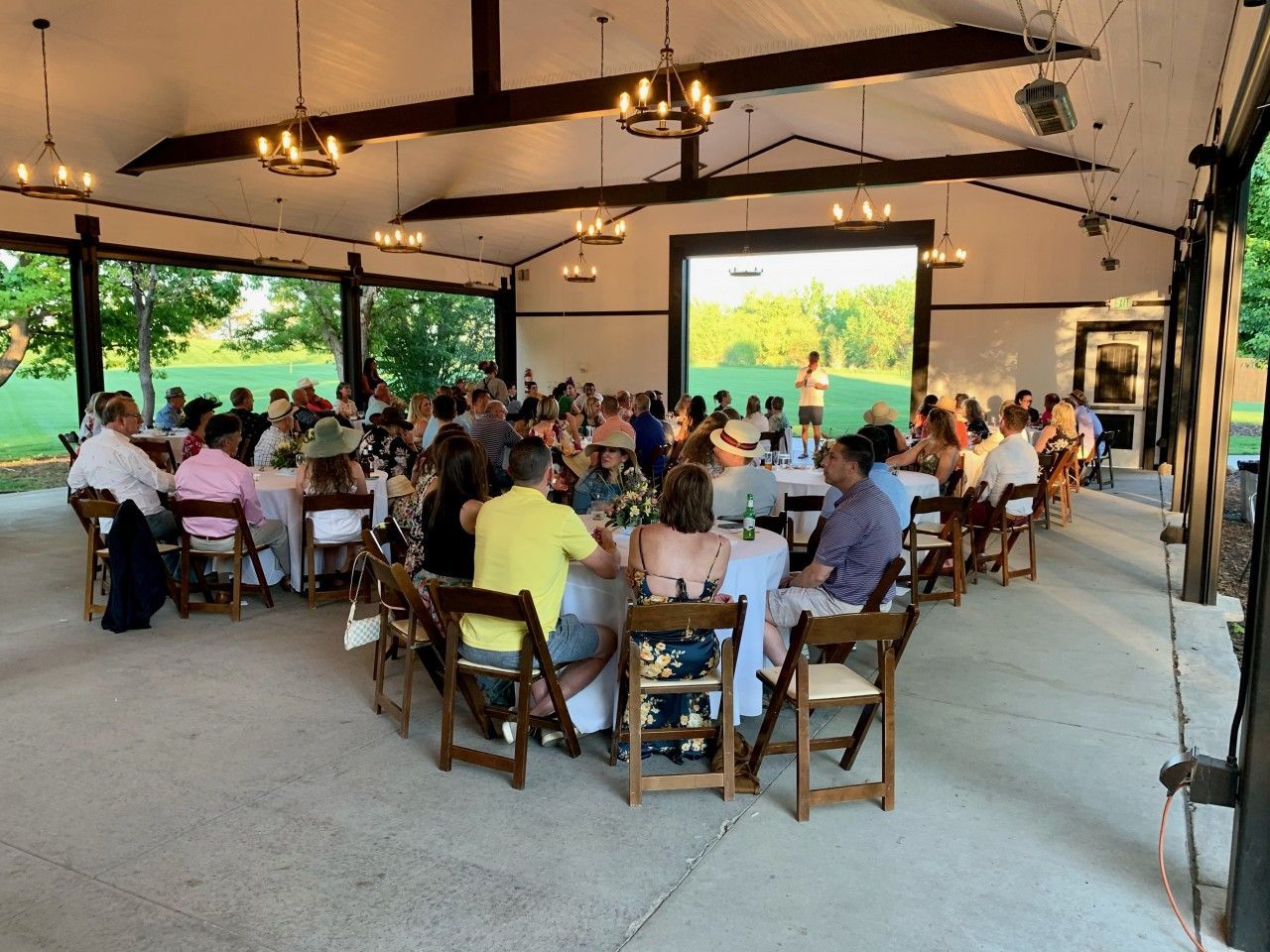 People seated at tables under a pavilion, likely at an outdoor event, with a screen in the background.