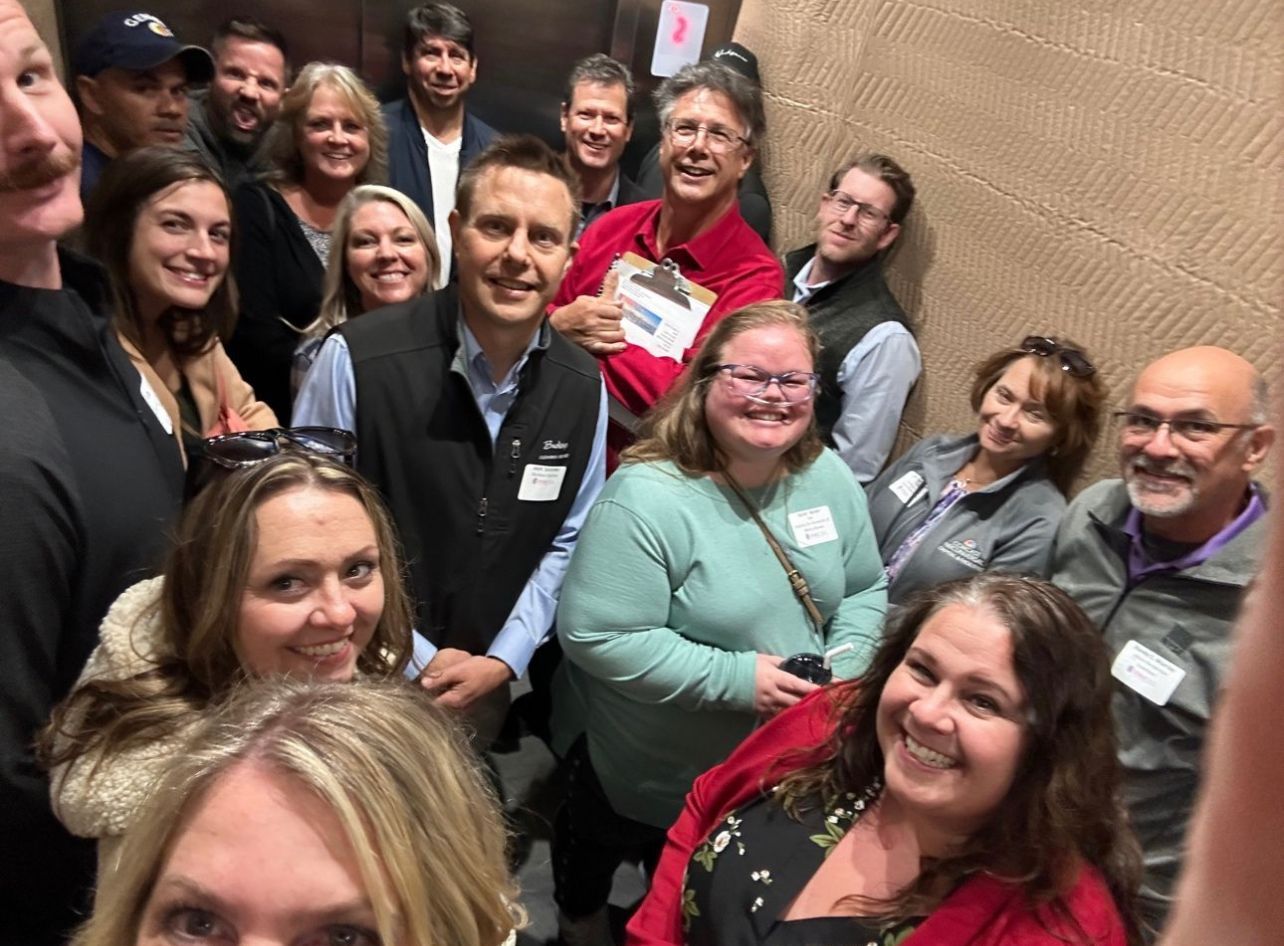 Group of people crammed in an elevator, smiling. Faces are close together.