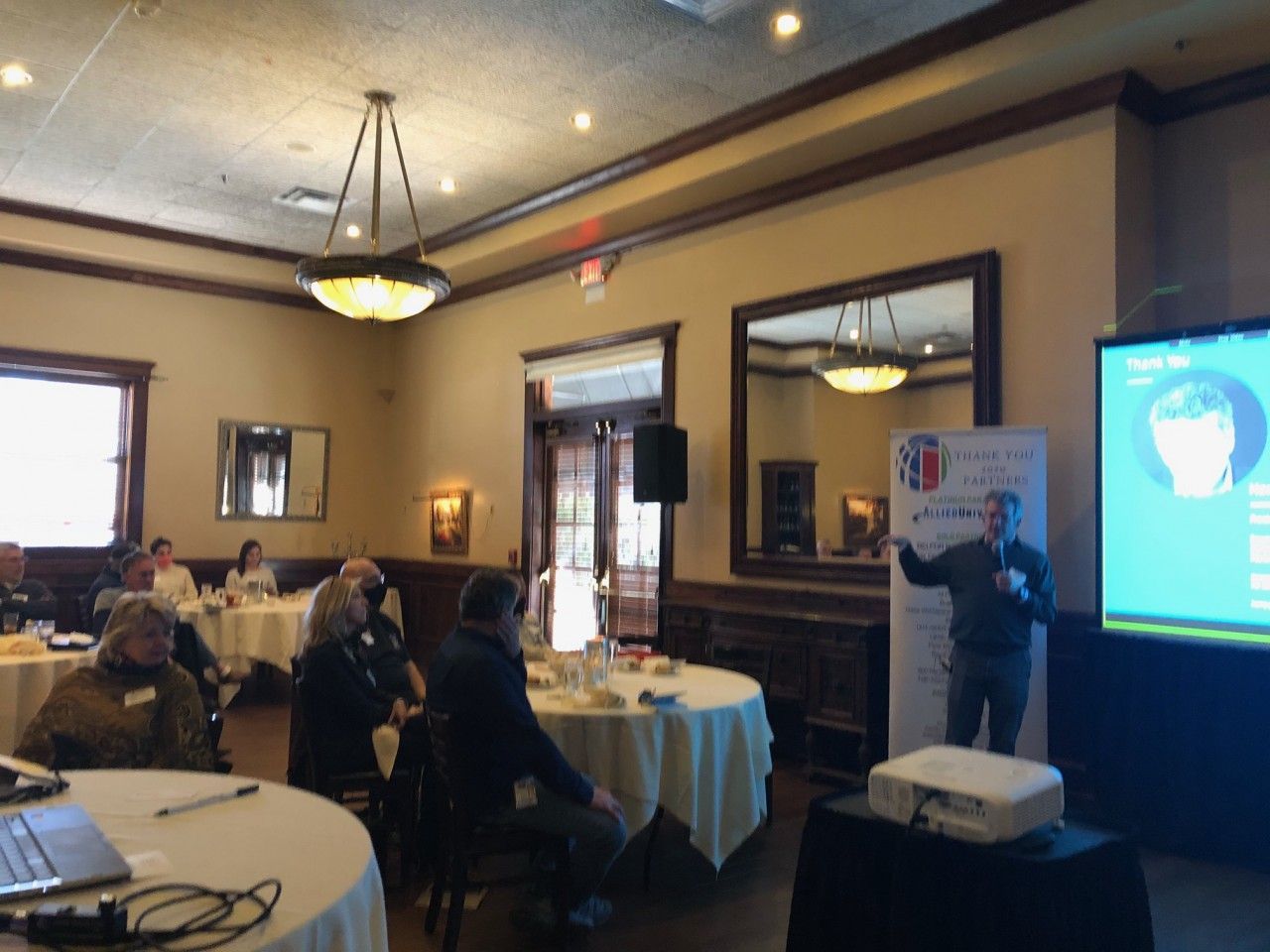 A man presents at a conference. Attendees sit at round tables. A projection screen and banner are visible.