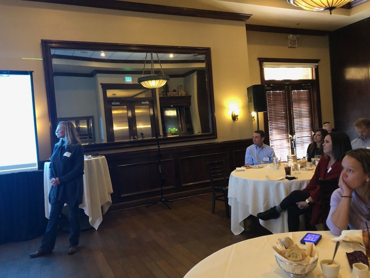 Woman presenting to a seated audience in a restaurant. White tablecloths, mirror, and a projector screen are visible.