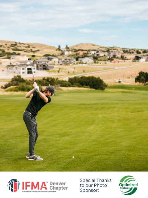 Golfer in a black shirt swings a club on a green course with houses in the background.