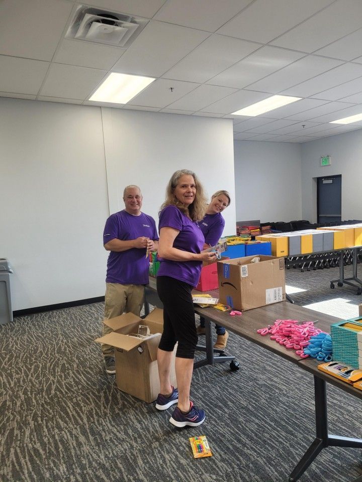 Three people in purple shirts sorting items at a table indoors.