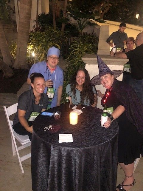 Group of people at an outdoor event, some dressed in scrubs and witch costume, seated around a table.