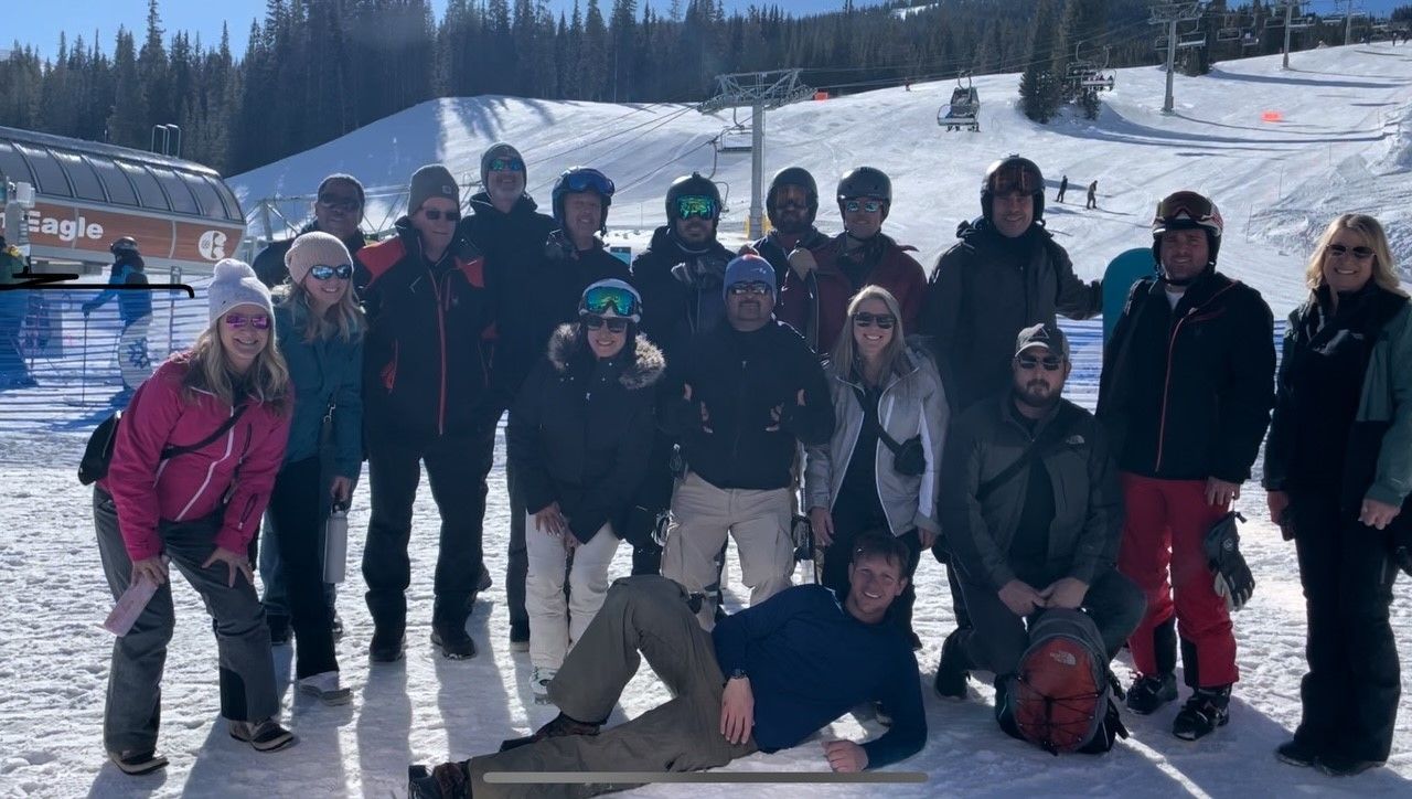 Group of people pose in snow, near ski lift. Some wear winter gear. Bright sunny day.