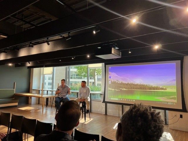 Two men sit on stools, speaking in front of a screen with a landscape. An audience observes.