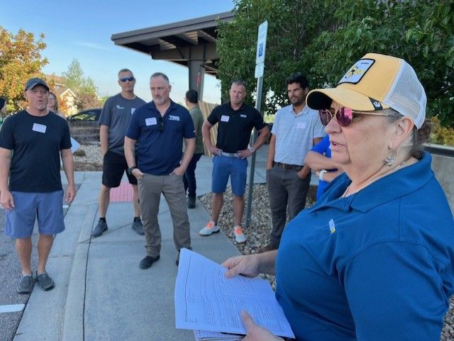 Group of people listen to a woman with papers. Outdoors by a building. Some wear name tags.