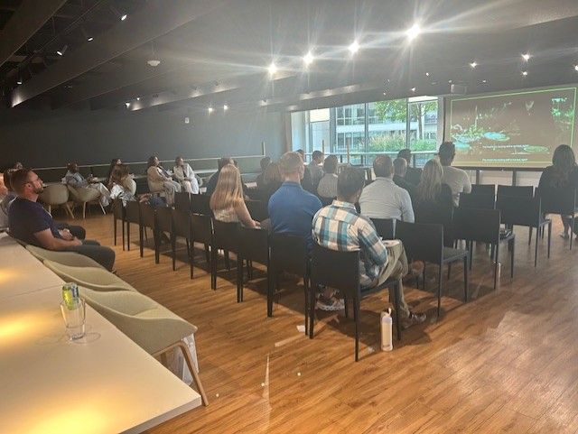 People seated in rows watching a screen during an event in a modern room with wood floors.
