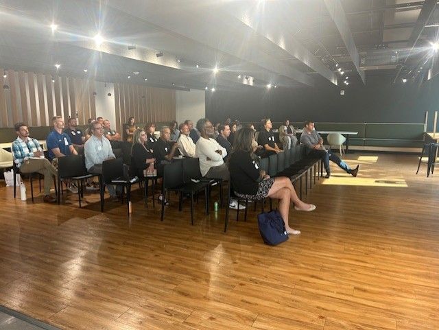 People in chairs at an indoor event, attending a presentation. Wooden floor, overhead lights, and a dark wall.