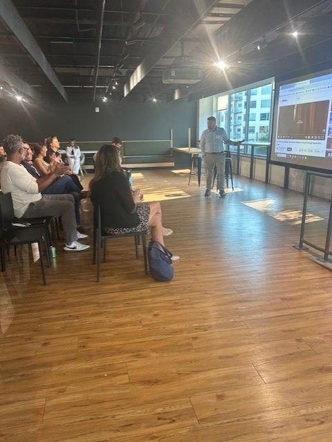 A person presents to a seated audience in a room with a screen and a large window.
