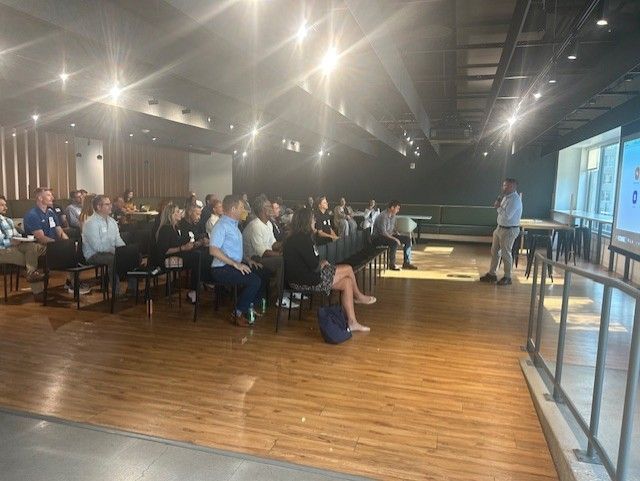 A presenter giving a talk in a room full of people. People sit in chairs and face the speaker.