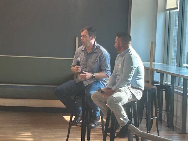 Two men seated on stools, one speaking into a microphone. They are indoors with a dark wall behind them.