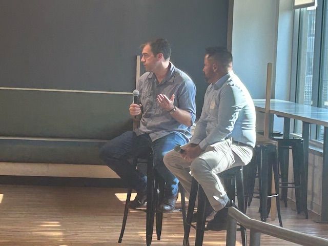 Two men sit on stools, one speaking into a microphone. They are in a well-lit room with a window and a bar-like area.