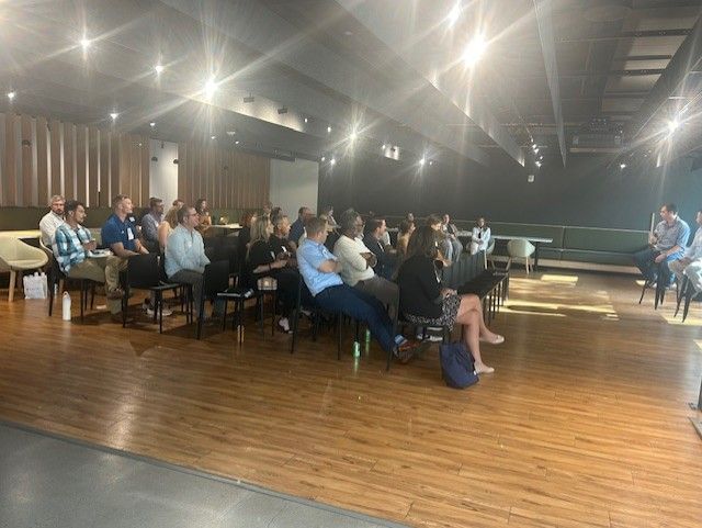Audience at a conference listens to a speaker in a room with wood floors and modern decor.