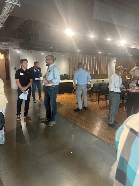People socializing in a well-lit event space with concrete floors, some stand near a wooden floor, and a door on the left.