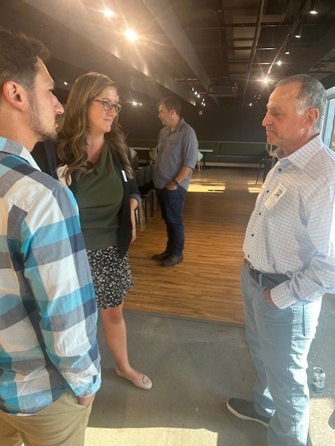 People in a meeting room, conversing. Woman in black cardigan, man in plaid shirt. Wooden floor, overhead lights.