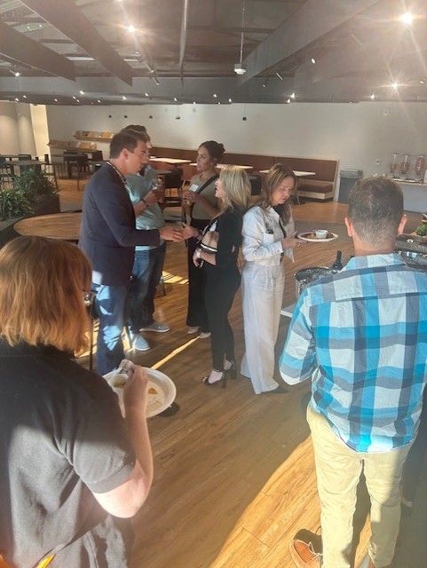 People socializing indoors, some holding food and drinks. Wooden floor, large windows, and neutral décor.