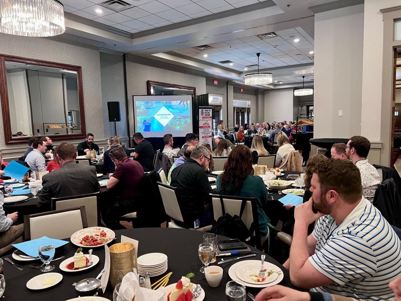 Conference attendees seated at tables, a presentation on screen, in a banquet hall.