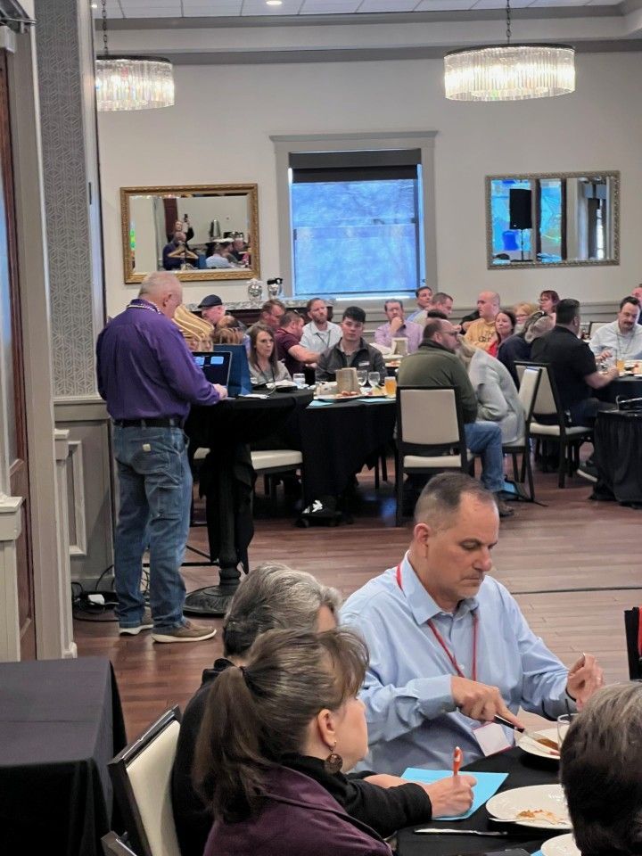 Man speaking at a conference. Attendees seated at tables, some eating. Formal setting, overhead lights.