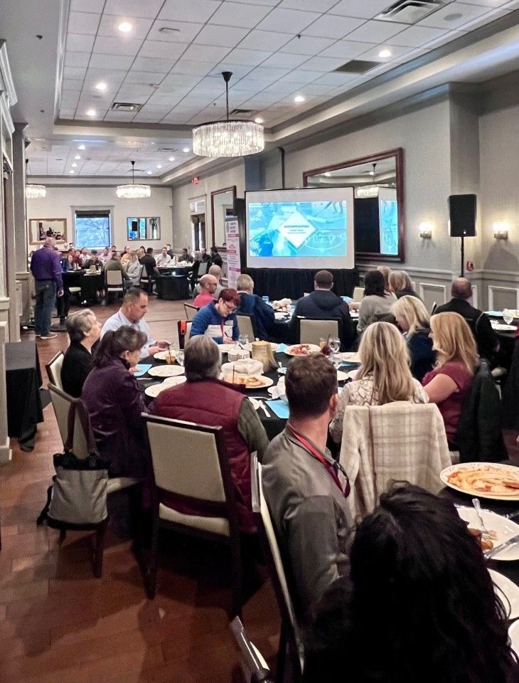 Conference room with attendees seated at round tables facing a projector screen displaying text.