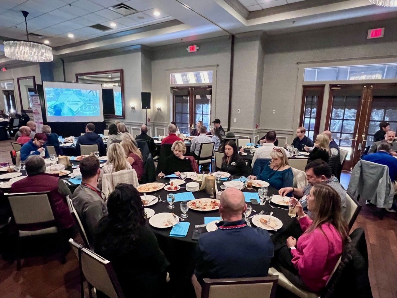 People at tables eating in a large room with a projector screen.