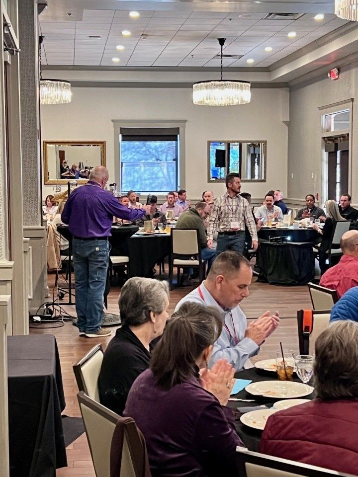 A man in purple shirt speaks at a conference in a dining room. People are seated at tables, clapping.